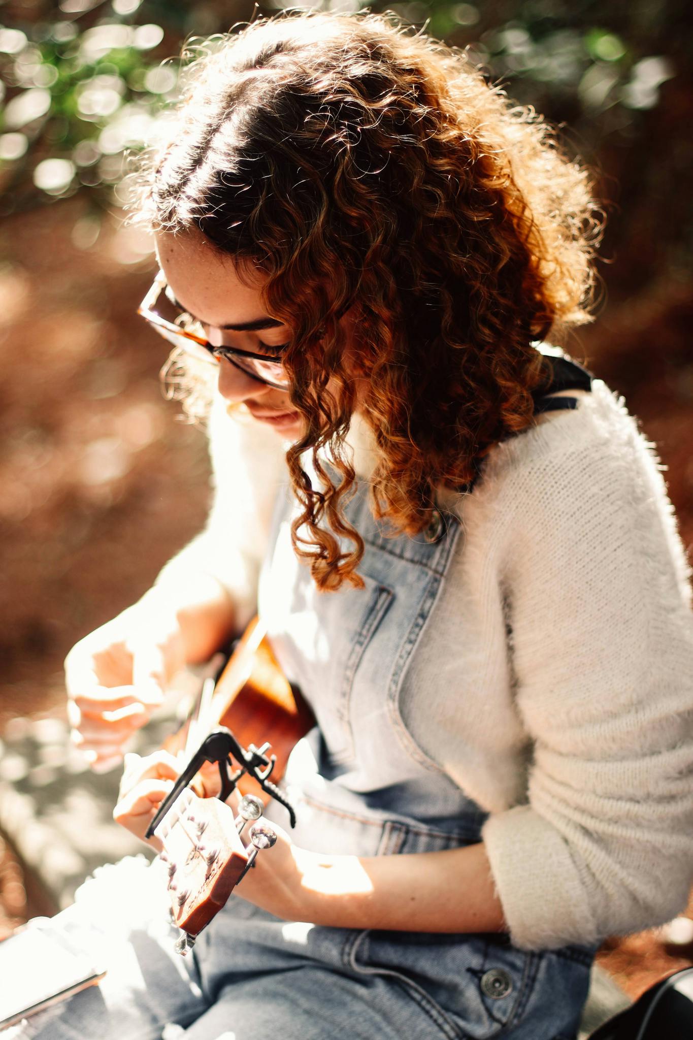 A woman sitting outdoors playing an acoustic guitar under sunlight, creating a warm, serene atmosphere.