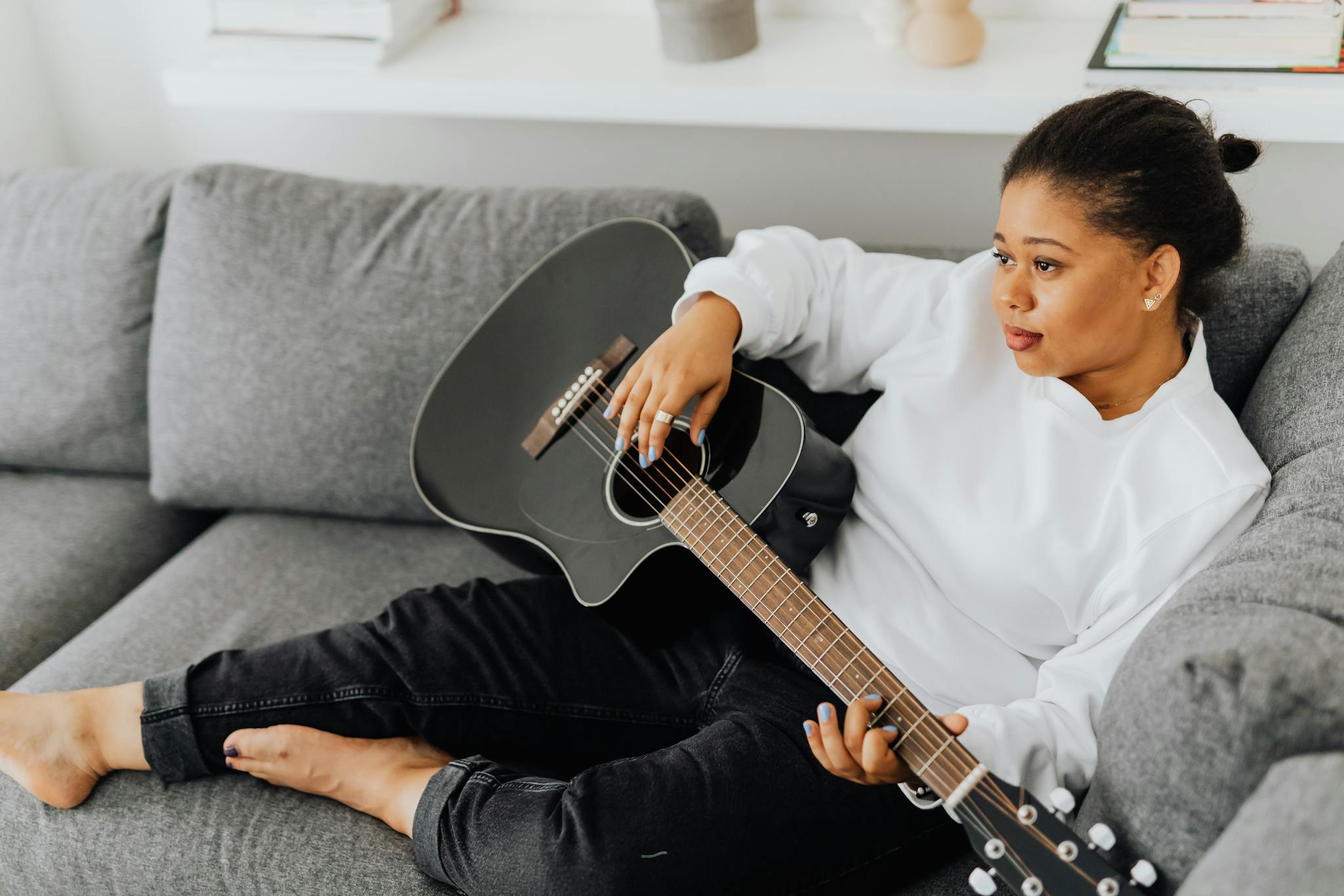Black woman in white long sleeves relaxing and playing an acoustic guitar on a comfortable gray sofa indoors.